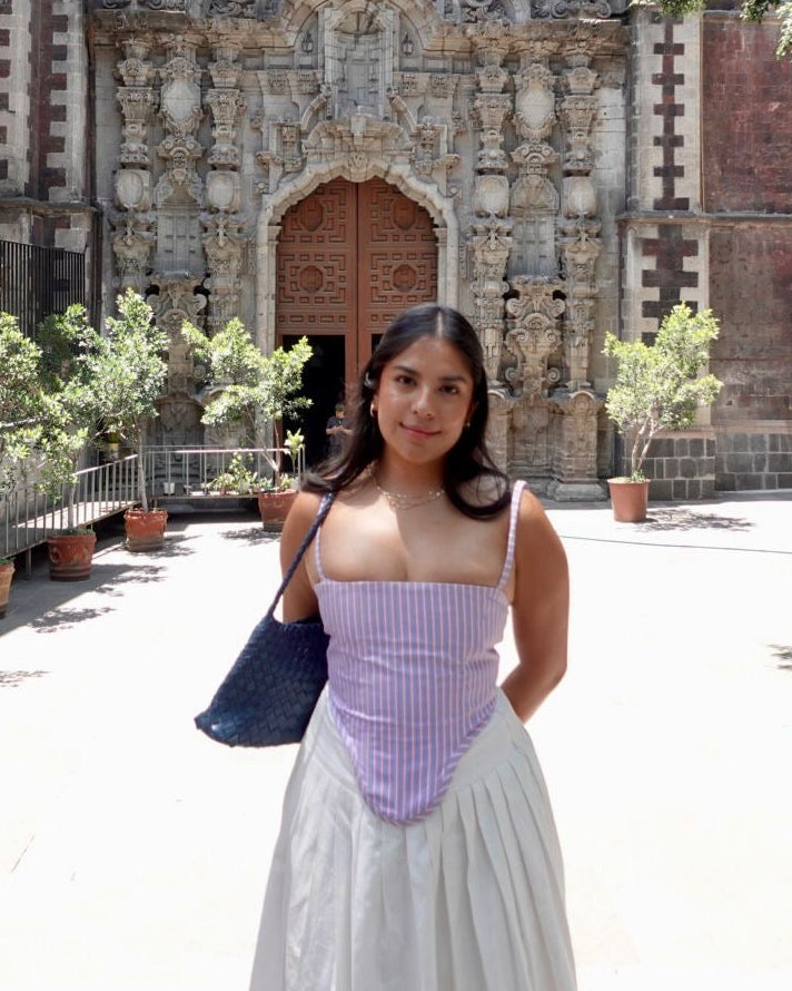 A woman wearing a lavender and white striped corset-style top with a long white skirt stands in front of an ornate historic stone church facade in a sunny outdoor plaza.